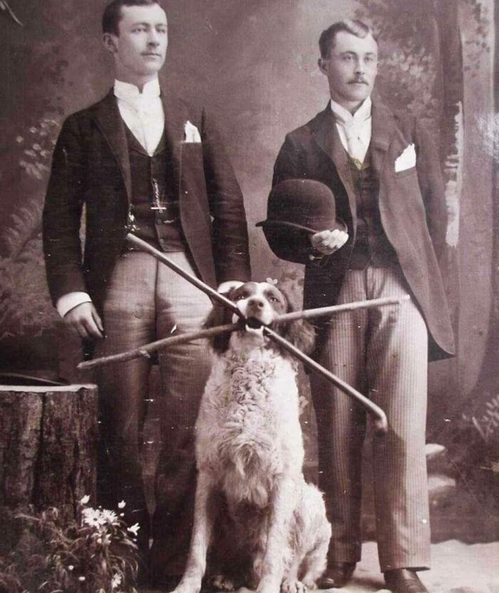Two formally dressed men pose with a dog holding sticks in its mouth, a charming historical photo from an earlier era.