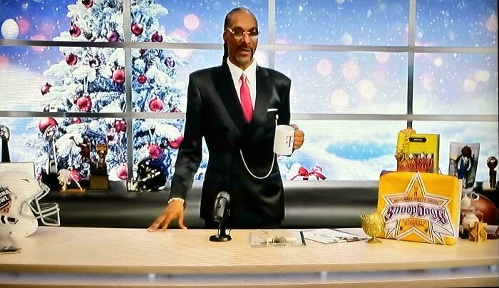 Snoop Dogg in a suit holding a mug behind a desk with a Christmas tree background, highlighting commercials people can't stand.