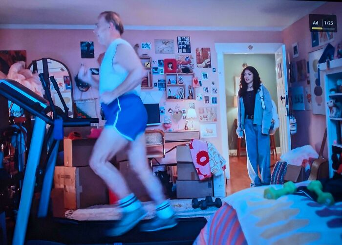 A man running on a treadmill in a cluttered room, a young woman watching in the background, a scene from commercials people can't stand.