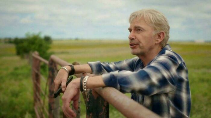 A man with gray hair leaning on a rustic fence, looking out over a vast field, a tranquil scene from commercials people can't stand.