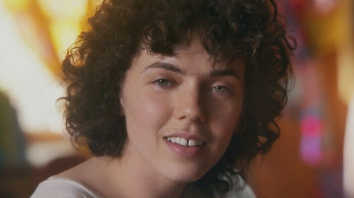 A young person with curly hair and a subtle smile, close-up shot, from a commercial.