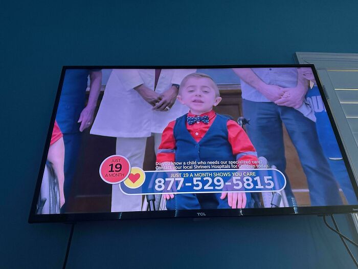 A young boy in a blue vest and bow tie looks at the camera with adults blurred behind him, a TV displaying a commercial with a call to action for donations to Shriner's Hospitals.