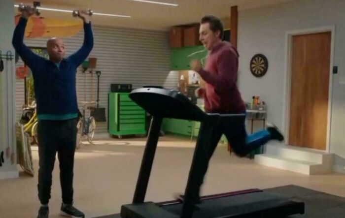 Two men exercising in a garage, one running on a treadmill and the other lifting weights, featuring people in commercials.