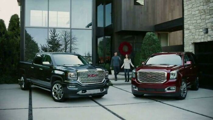 Two GMC vehicles, a dark pickup truck and a red SUV, parked in front of a modern home, commercials people cant stand.