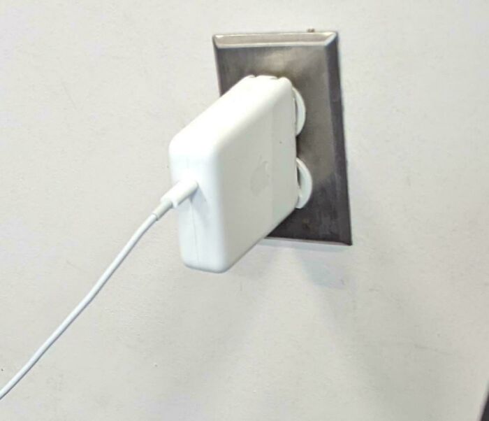 An Apple charger barely plugged into an outlet, hanging precariously, reflecting behavior of people who think they're the main character.