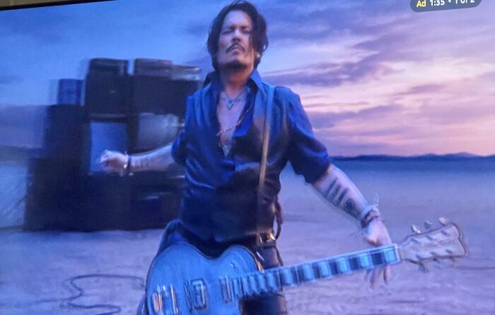 A man with a mustache and tattoos on his arms, wearing a blue shirt and vest, holding a guitar in a commercial.