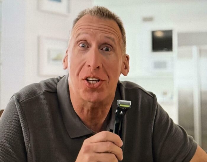 A man with short brown hair holds a black razor, looking directly at the camera with a surprised and slightly open-mouthed expression in a commercial.