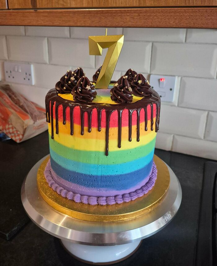 A vibrant rainbow cake with chocolate drip and a gold number 7 candle, an amazing treat from bakers.