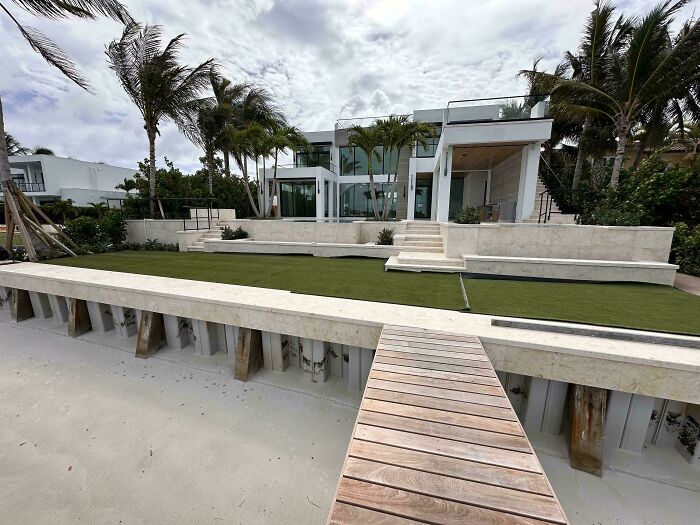 Modern architecture design of a waterfront home with palm trees and a private pier leading to a sandy beach.