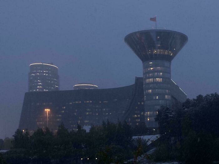 Illuminated modern architecture designs skyscraper with a distinctive conical top, rising above trees under a dark, cloudy sky.