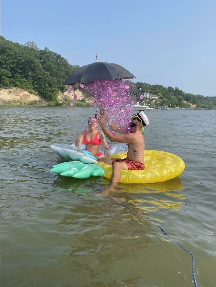 A couple on inflatable rafts in water during a gender reveal moment with pink confetti from an umbrella.