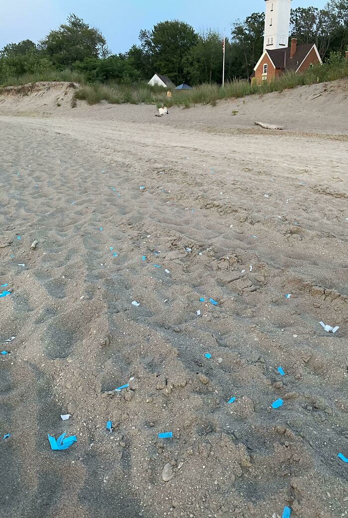 Blue confetti and litter scattered across a sandy beach from a gender reveal event, near a lighthouse.