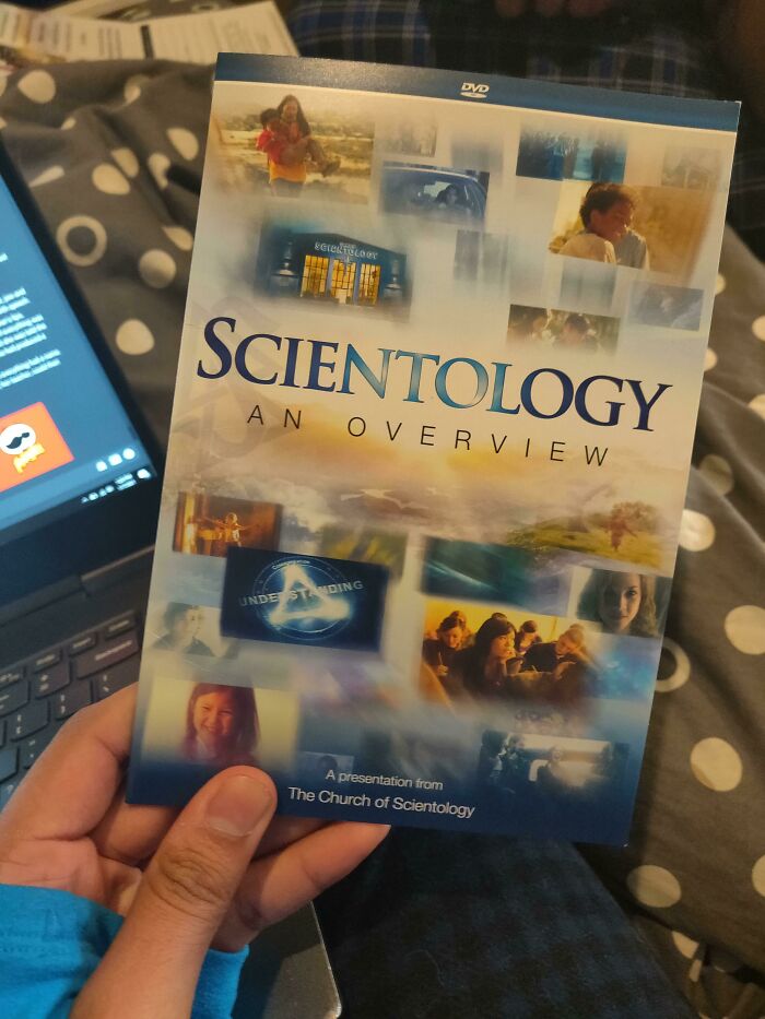 A hand holds a Scientology DVD, an overview, freaked out a man, lying on a patterned blanket in a woman's home.