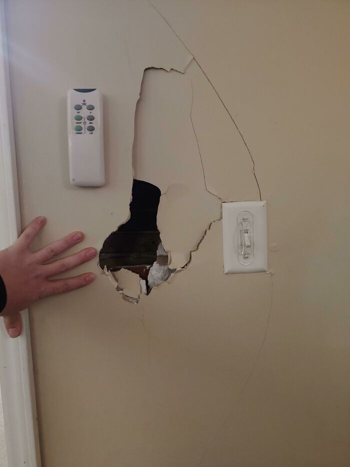 A large hole freaked out a man in a wall near a light switch and remote control in a woman's home.