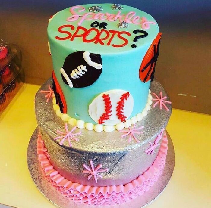 A two-tier gender reveal cake decorated with Sparkles or Sports theme, featuring a football, basketball, and baseball.