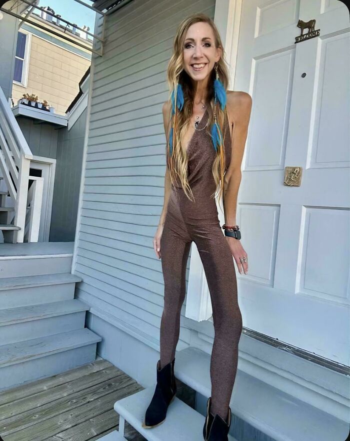 A woman in a brown shimmery jumpsuit and blue feather earrings posing on steps, trying to look natural on social media.