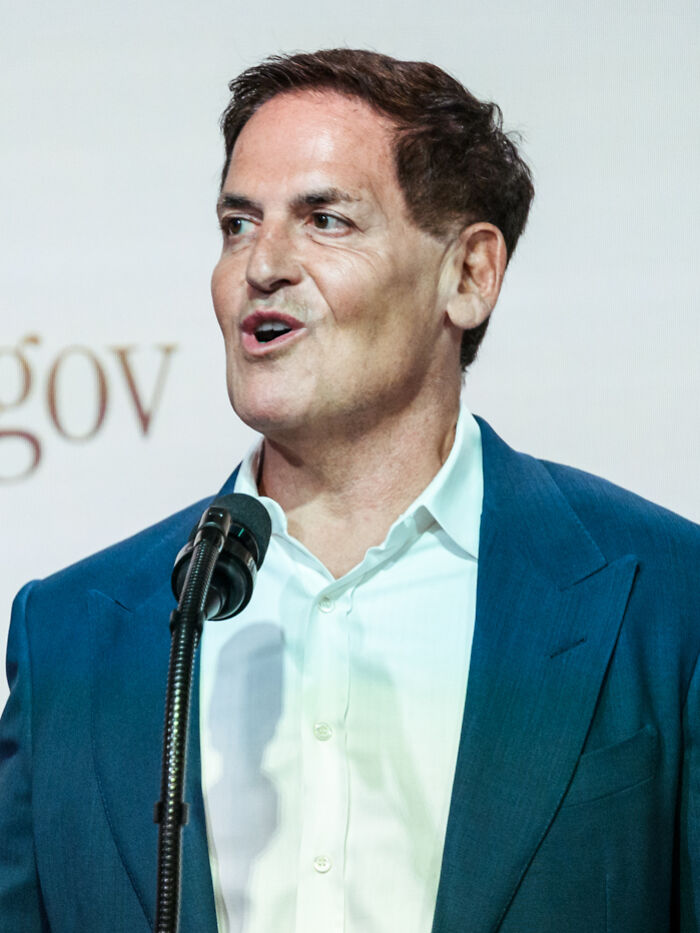 Mark Cuban speaking at a microphone, showcasing his prominent career highlights and entrepreneurial journey.