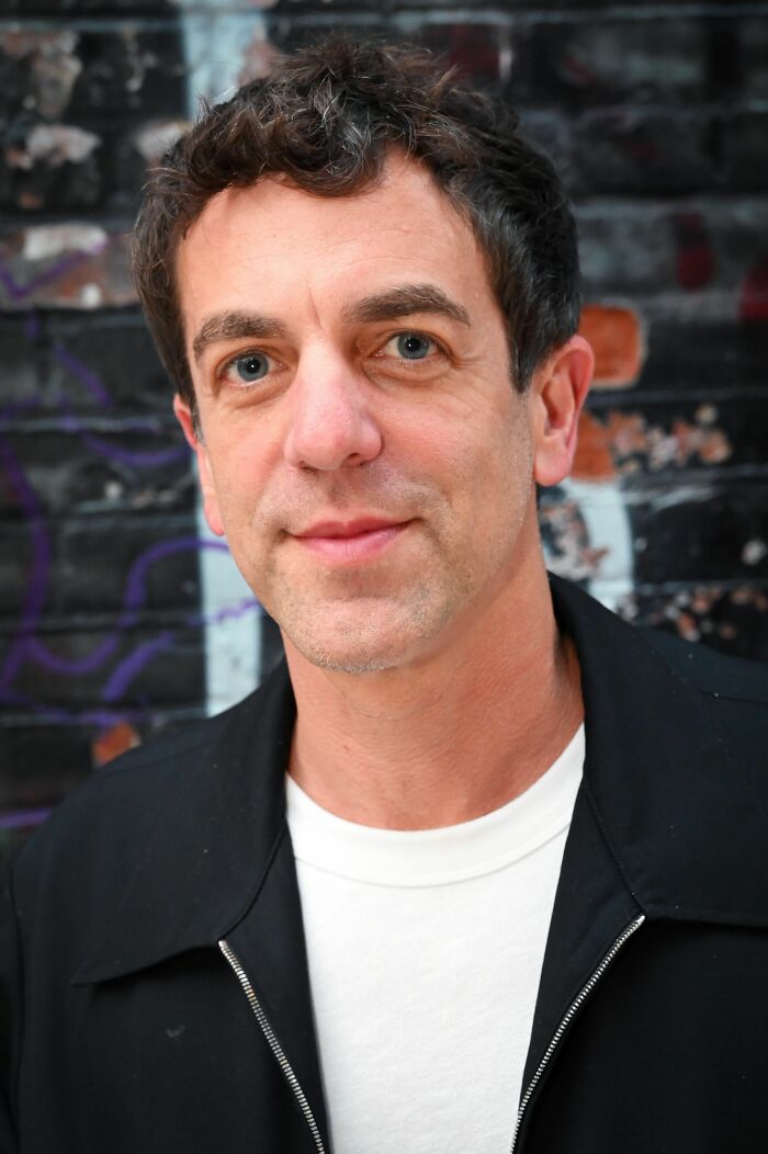 B. J. Novak smiling gently in a close-up portrait, standing against a blurred brick wall, showcasing his career highlights.
