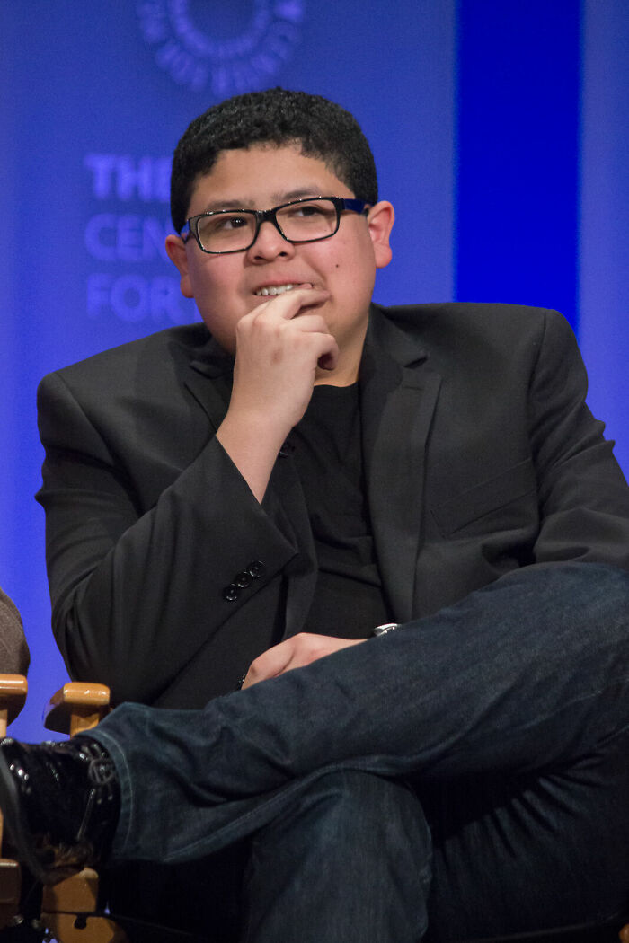 Rico Rodriguez, wearing glasses and a black suit, sits with his hand to his chin, contemplating during a career highlight event.