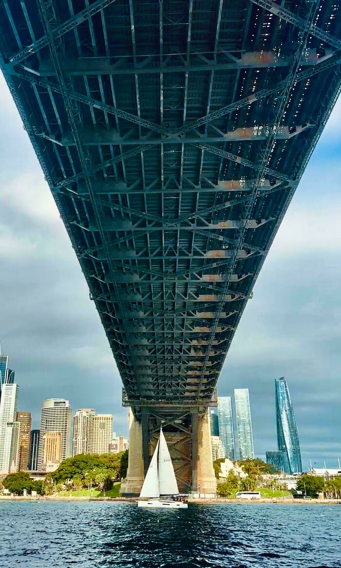 A majestic view looking up at the Sydney Harbour Bridge with a sailboat passing underneath, an interesting post for Aussies.