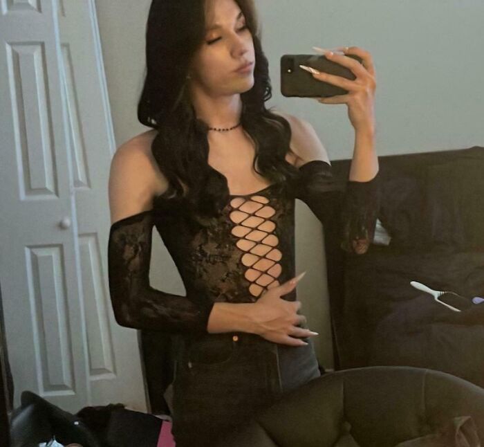 A person with amazing long hair posing for a mirror selfie indoors, wearing a lace-up top and dark pants.