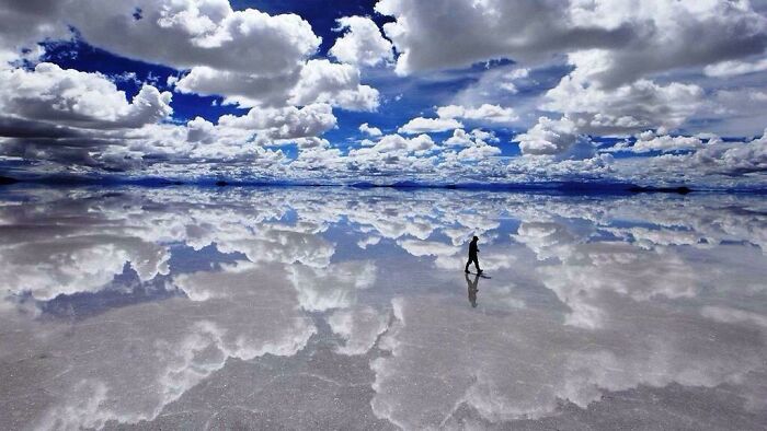 A person walks across a vast salt flat, perfectly reflecting the clouds above, a real photo that makes your brain question reality.