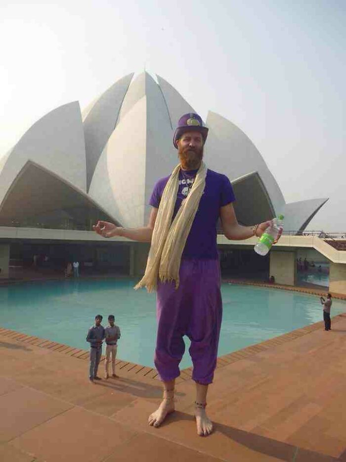 A man in a purple outfit posing in front of a lotus-shaped building, a real photo that makes your brain question reality.