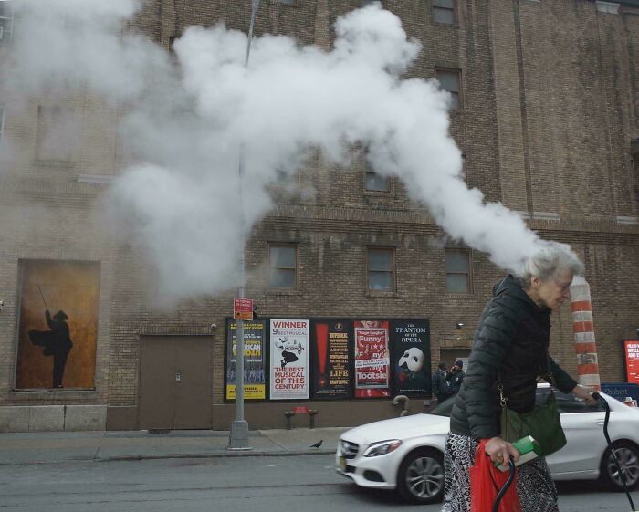 An older woman walking past a building with steam emerging from a pipe that appears to be coming from her head, a real photo that questions editing.