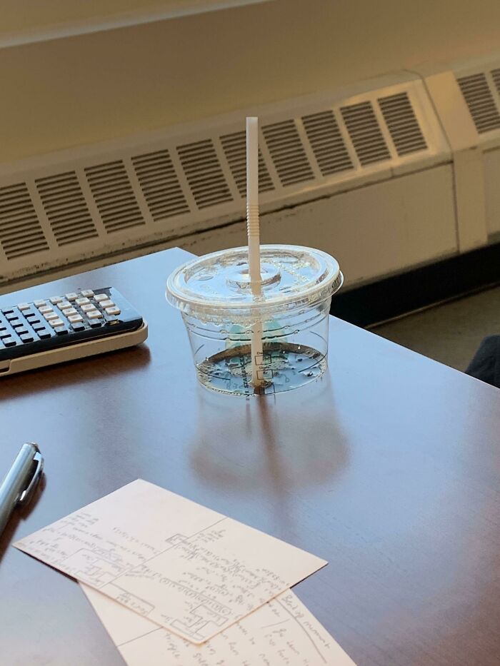 A clear plastic cup with a straw sits on a wooden desk, containing a small amount of liquid, a real photo that makes your brain question if it was edited.