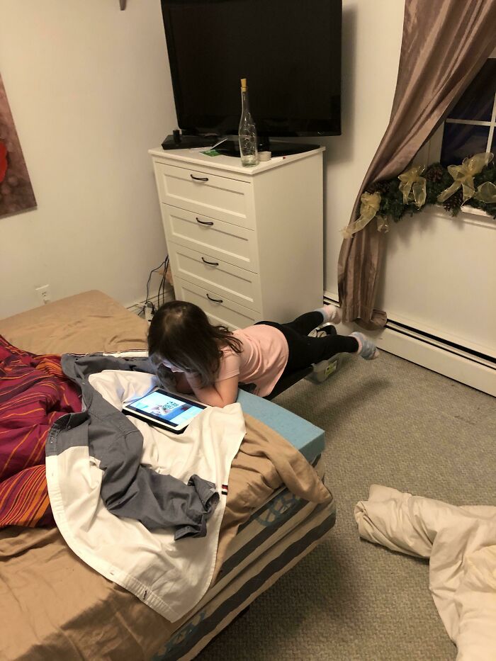 A young girl lies face down on a small blue stool looking at a tablet, showcasing a real photo that makes your brain question if it was edited.