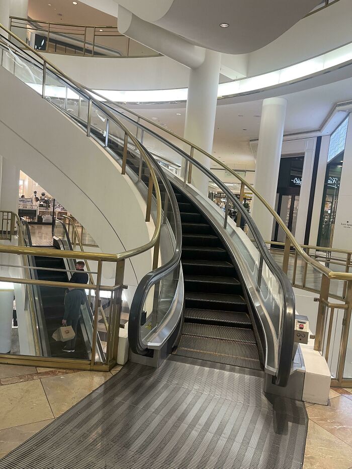 A curved escalator with golden railings in a modern shopping mall, real photos that make your brain question if they were edited.
