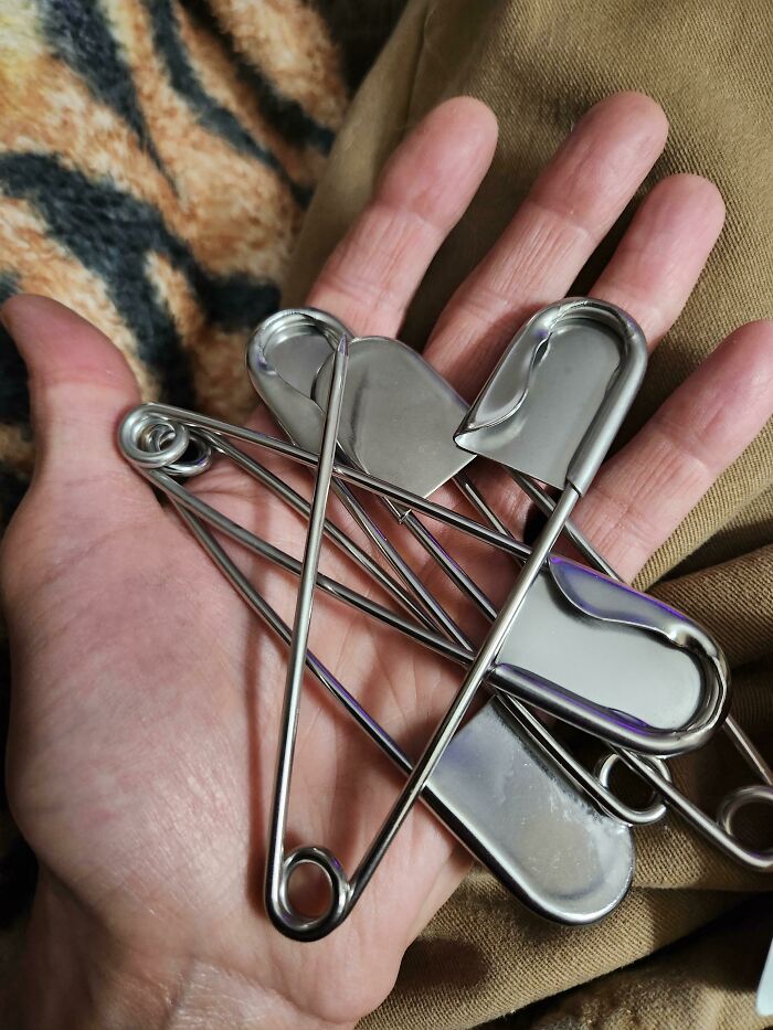 A hand holding several large safety pins, making the brain question if the photo was edited due to unusual perspective.