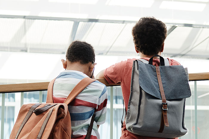 Two kids with backpacks, seen from behind, leaning against a railing, possibly discussing unfortunate names.