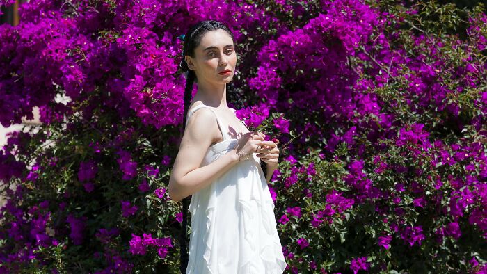 A woman in a white dress standing in front of purple flowers, looking thoughtful about not wearing a boyfriend's gift.
