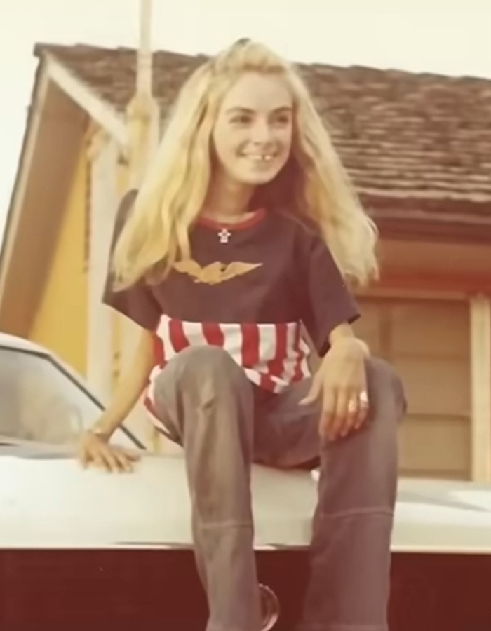 A smiling young polio survivor with long blonde hair sits on a car, wearing an American flag shirt, living in an iron lung.