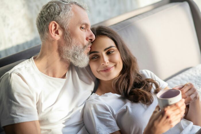 A happy age-gap relationship couple, an older man and younger woman, relaxing in bed.