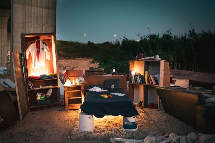 An outdoor bedroom setup under a bridge with a bed, shelves, and clothes, illuminated at night. reFocus Contest photo capturing the world.