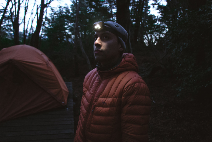 A person in a winter jacket and headlamp standing near a tent in a dark, creepy forest, highlighting mysterious things.