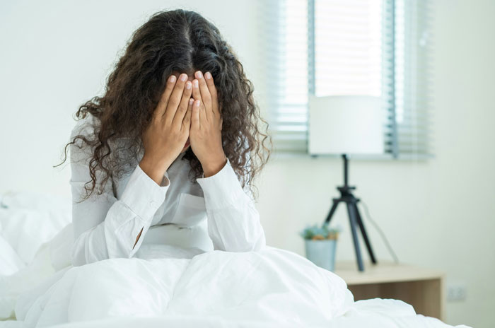 A woman with curly hair sits on a bed, hiding her face in her hands, reflecting men's complaints.