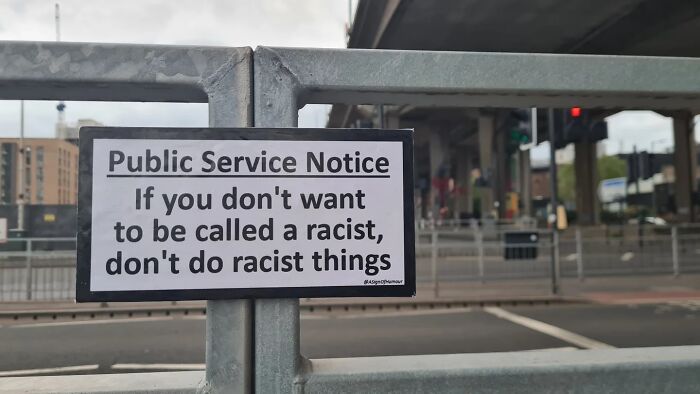 Absurd sign on a metal railing, stating a public service notice about not doing racist things.