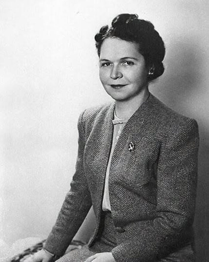 A black and white portrait of an overlooked woman in history, wearing a tweed suit and a small brooch, looking directly at the camera with a subtle smile.