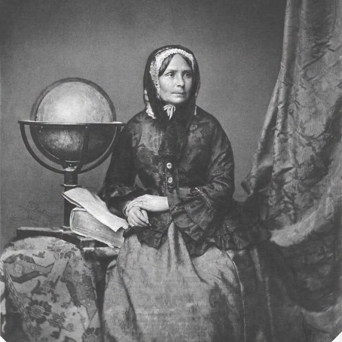 A black and white portrait of an overlooked woman in history, wearing a bonnet and a dark dress, seated next to a globe and books, looking to the side.