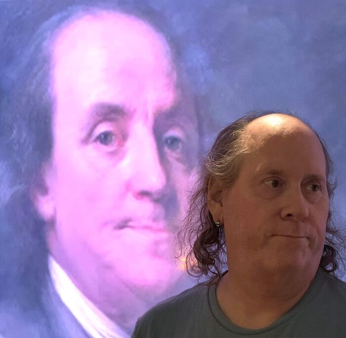 A man with long hair looking to the right, with an image of Benjamin Franklin in the background. His look may not match surprising facts.