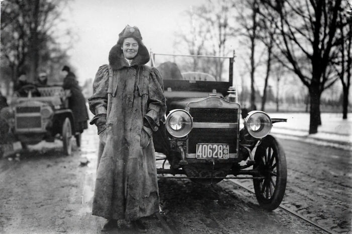 A black and white photo of a woman in a fur coat standing next to an early automobile, showcasing women overlooked in history.