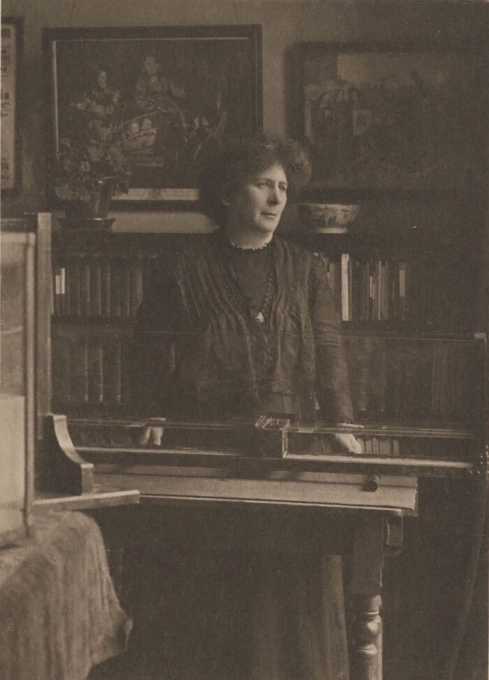 A sepia-toned image of an overlooked woman in history standing at a desk, looking to the side.