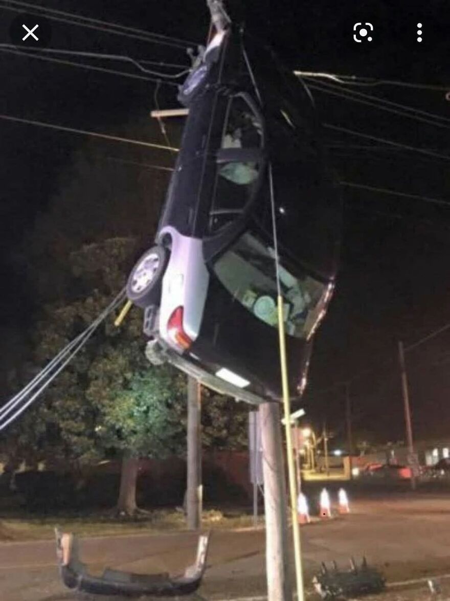 A dark car is suspended vertically from power lines after a rough day behind the wheel, defying gravity.