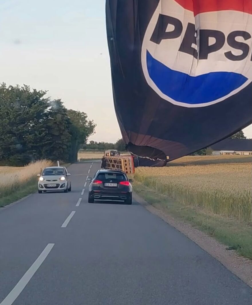 A large Pepsi hot air balloon landed near a road with cars, a rough day behind the wheel.