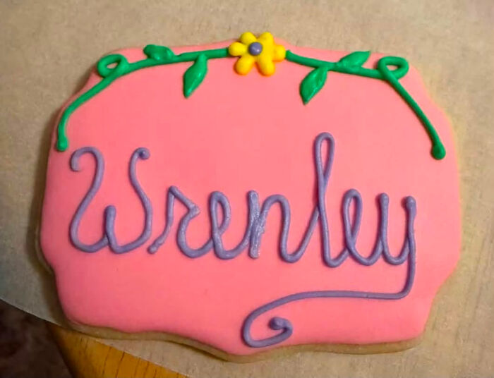 A pink rectangular cookie with a purple iced baby name Wrenley, adorned with green vines and a yellow flower.