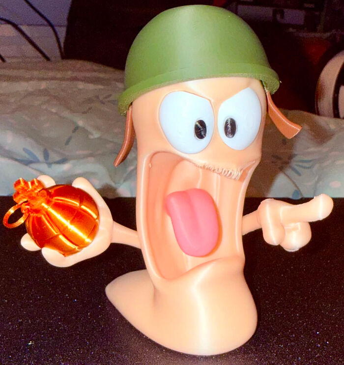 A 3D printed figurine of a cartoon worm in a military helmet, holding a glowing b**b and pointing, with an open mouth and tongue out.