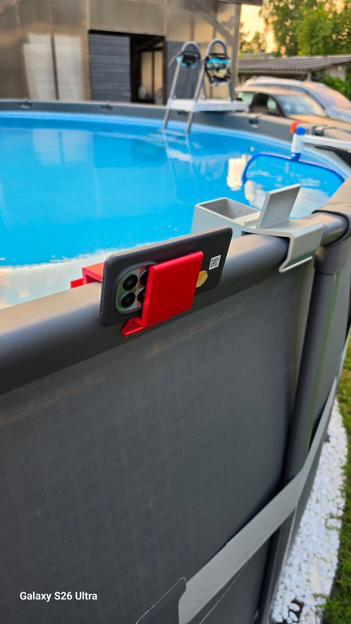 A smartphone in a red 3D printed holder attached to the edge of a swimming pool, a cool and useful item.